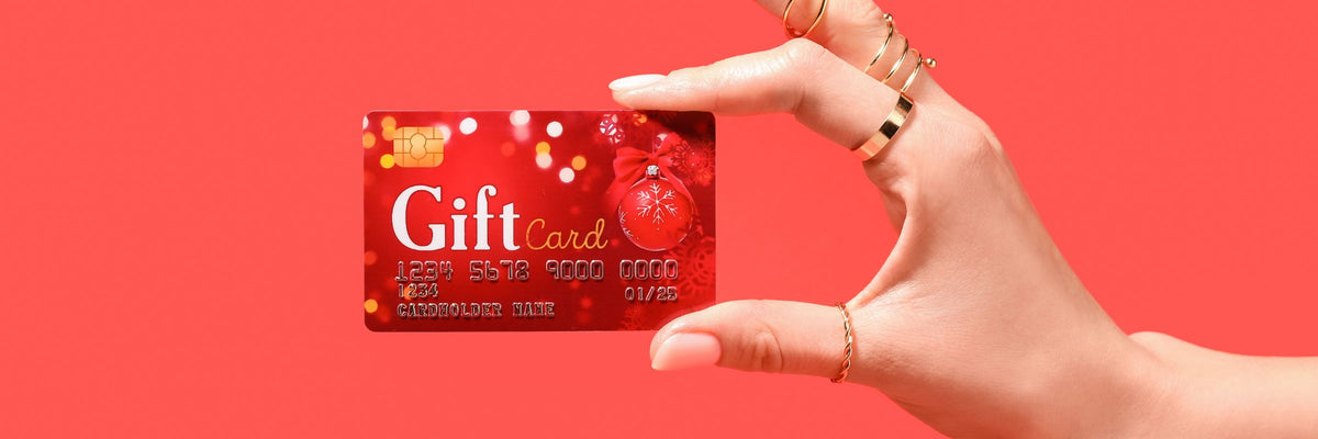 Une main aux bagues dorées tient une carte cadeau rouge sur fond corail, la carte cadeau Cosmesud